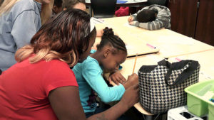 A third grade student sits and writes hers story as a family member looks over her shoulder.