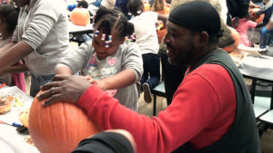 A kindergarten student cleans out her pumpkin while her helper watches over her.