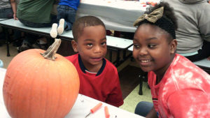 A kindergarten student and his middle school helper pose for a picture while they work on their pumpkin.