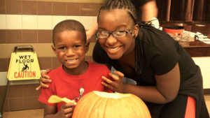 A kindergarten student and his middle school helper pose for a picture while they work on their pumpkin.