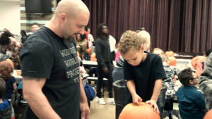 A kindergarten student works hard to clean out his pumpkin while his helper watches over him.