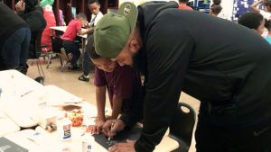 A 1st grade student watches on as his guest helps with their craft for family day.