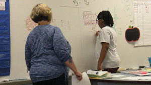 A student works on a math problem at the board while her teacher looks on.