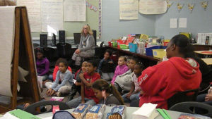 Students and family members sit at the carpet area while the teacher explains an activity.