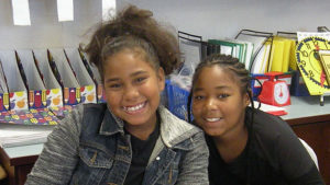 Two students smile for the camera while working at their desks.