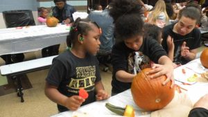 A student looks on as her helper scoops out their pumpkin.