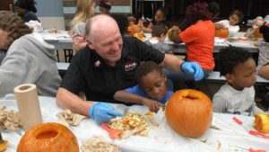 A helper looks on as the student shows where to carve the pumpkin.