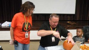 Miss Mel looks on as Mr. Bell helps a student carve his pumpkin.