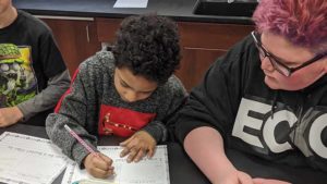 An Eighth grader watches on and helps a Second grader with his assignment.
