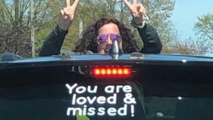 Ms. K peeks out above her car with a sign that says "You are loved and missed".