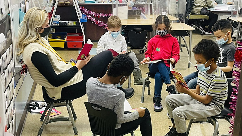 A group of first graders sit with their teacher in a circle while reading through a book.