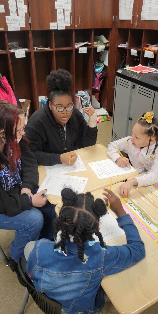 Eighth grade students sit down with a group of first graders to help them with their assignment.