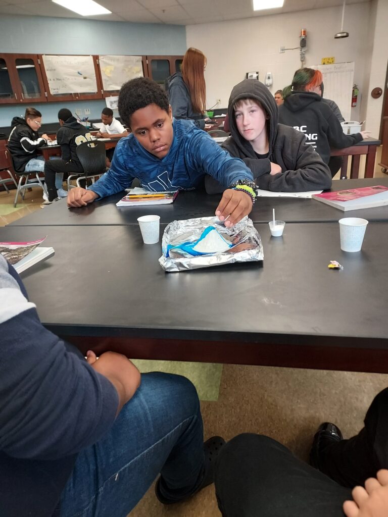 A student pours the liquid on his experiment as others watch.