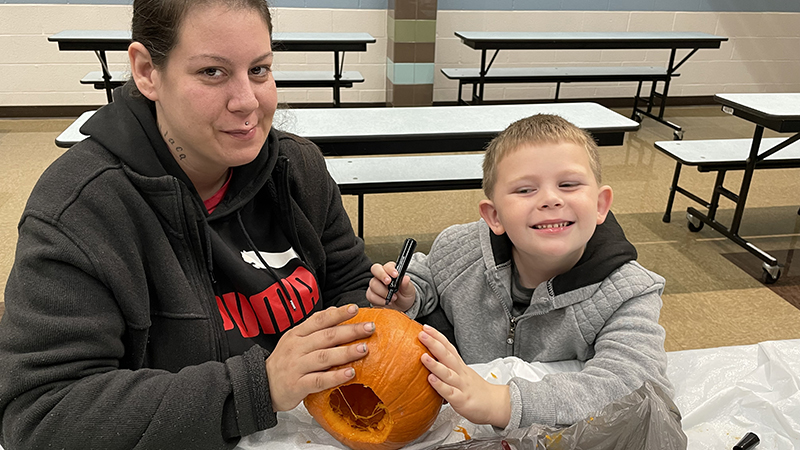 A student and his helper use a sharpie to draw on his pumpkin.