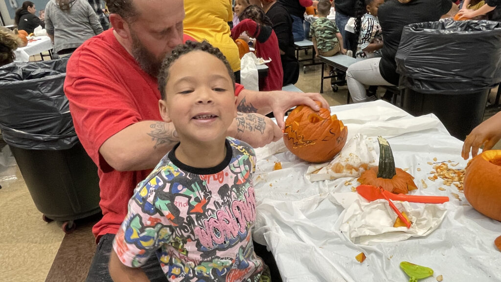 A student stops for a smile as his helper carves the pumpkin.