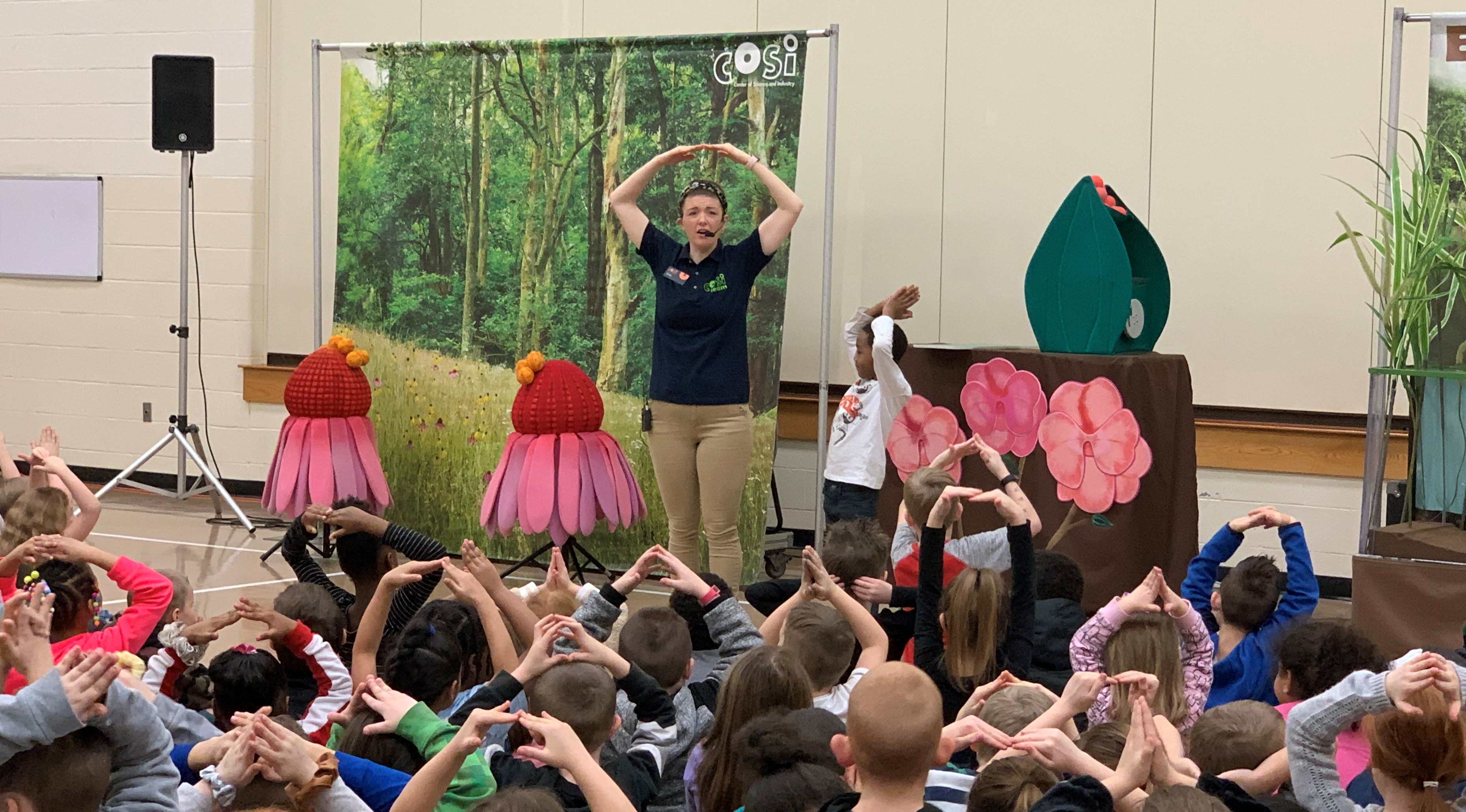 Students in a gym setting sitting on the floor watching a science lesson put on by COSI